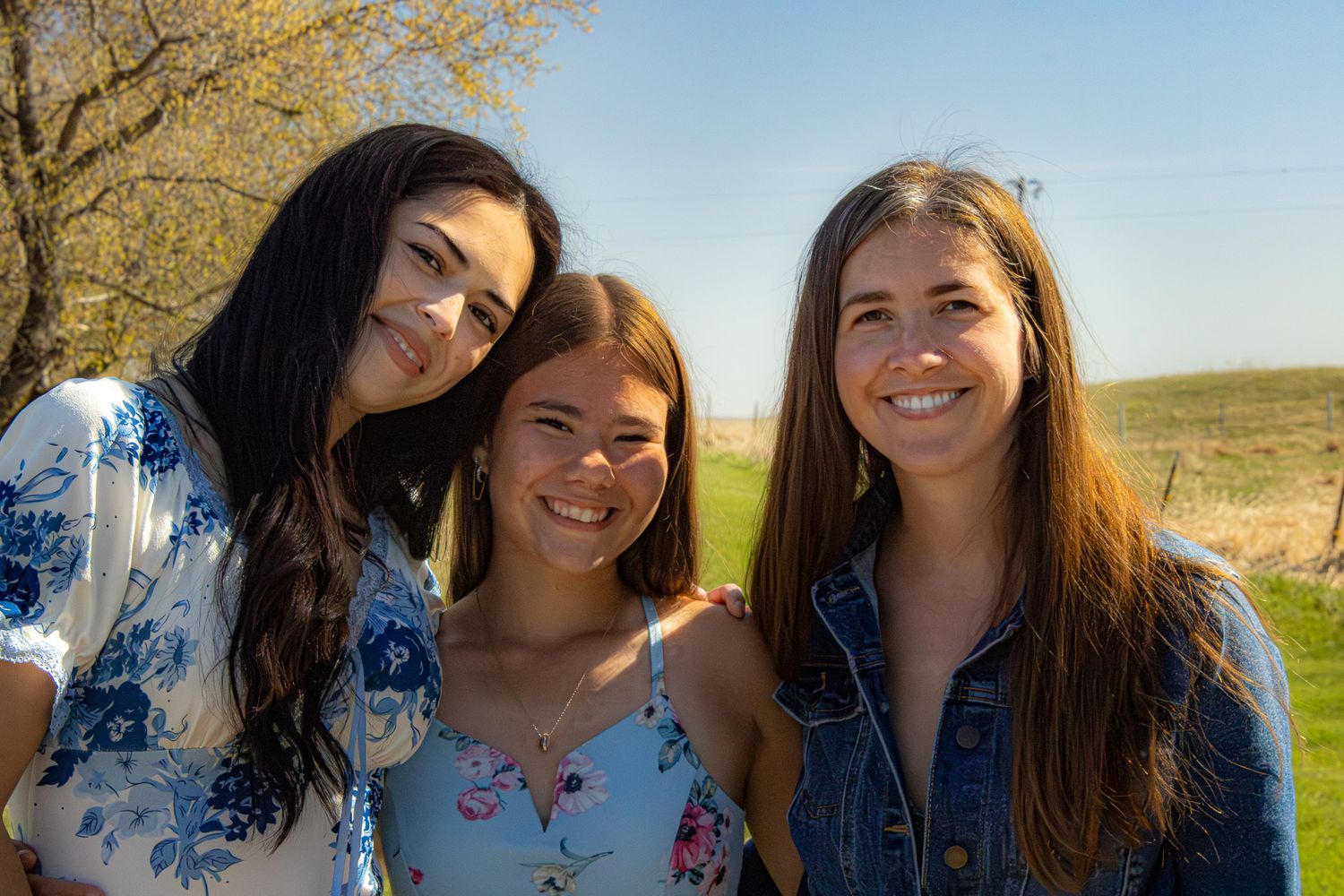 Three young women smile together outdoors on a sunny day with green fields in the background.