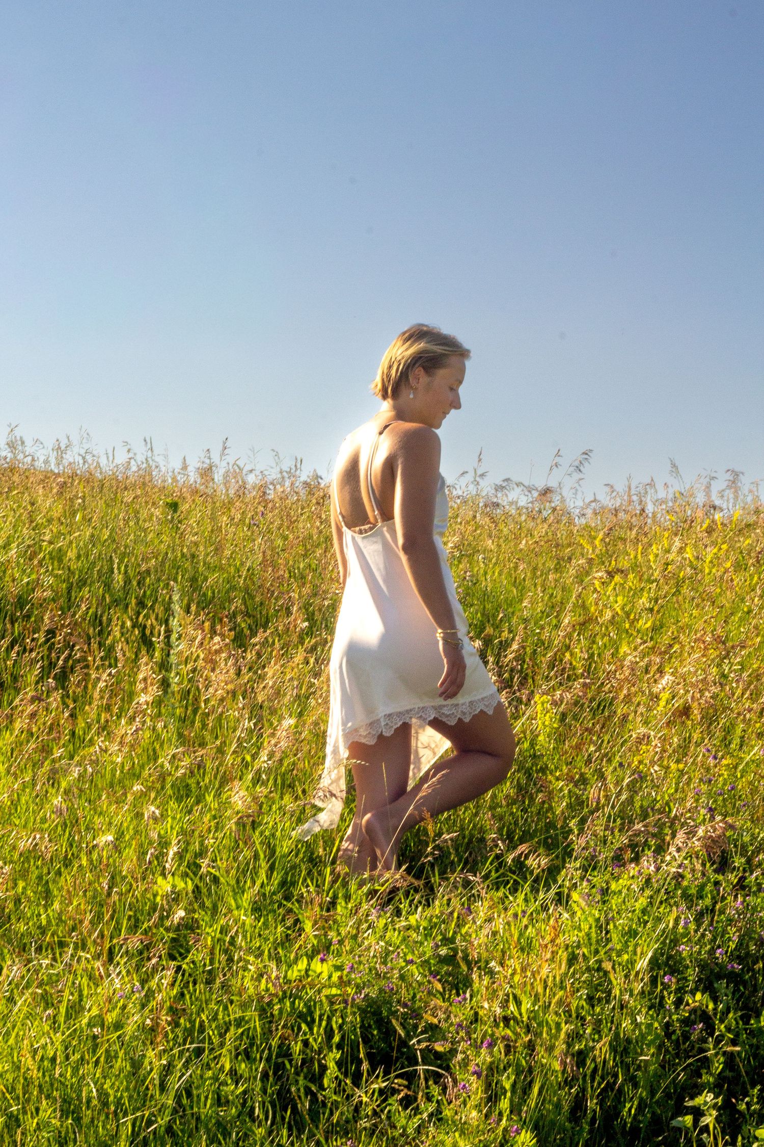 Young blonde woman in white sundress walking through lush green meadow under clear blue sky on sunny day.