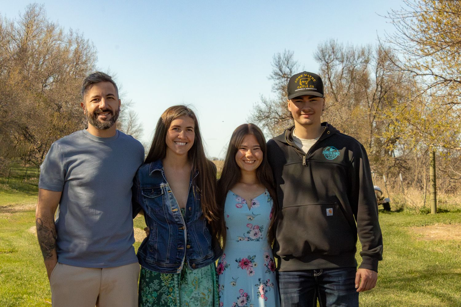 Four smiling people posing together outdoors in a park with green trees on a sunny day.