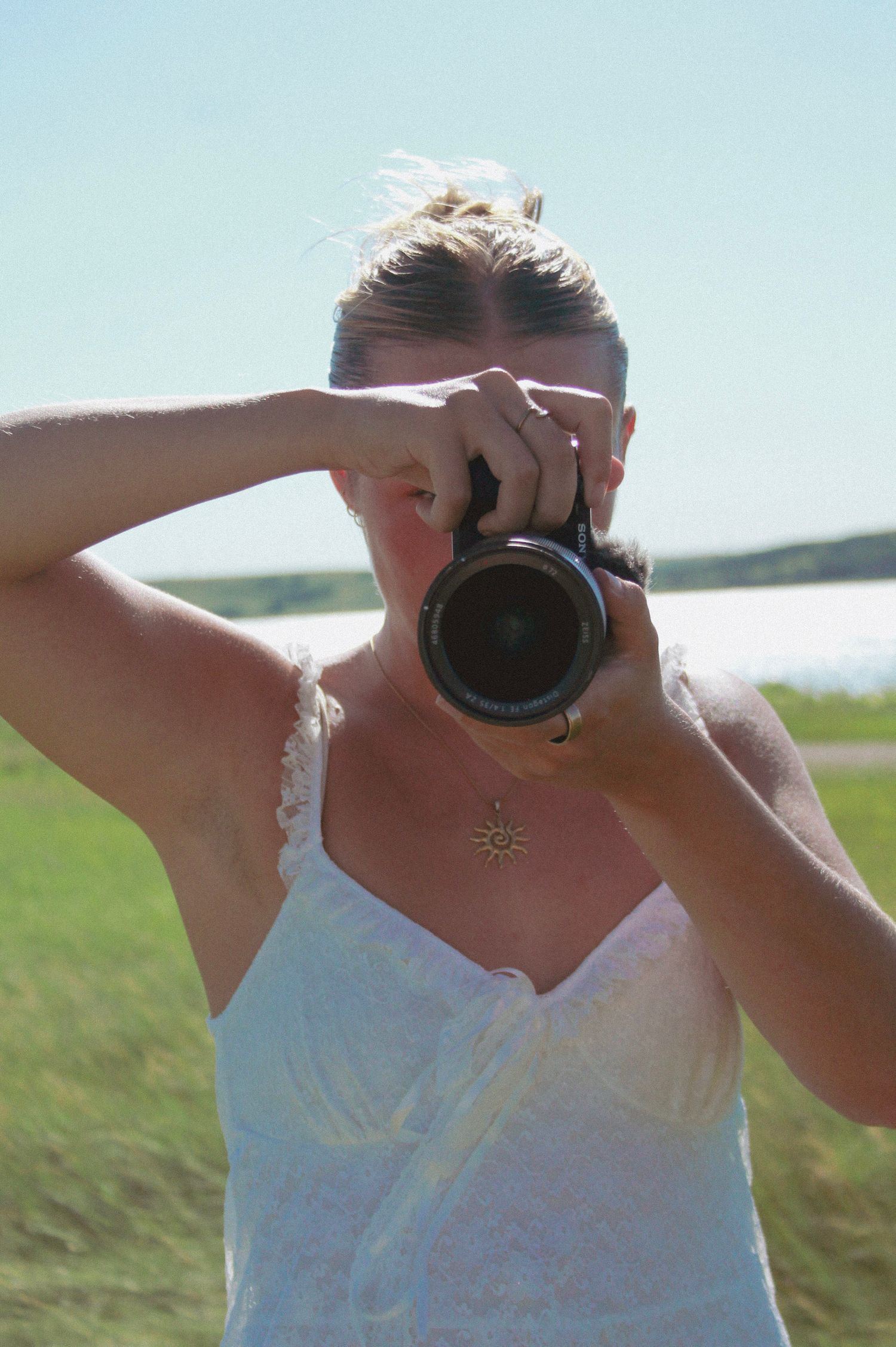 A photographer takes pictures with a DSLR camera while wearing a white summer dress outdoors.