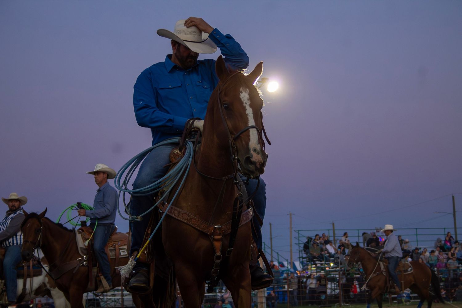 Cowboys compete in a nighttime rodeo event under bright arena lights while spectators watch from the stands.