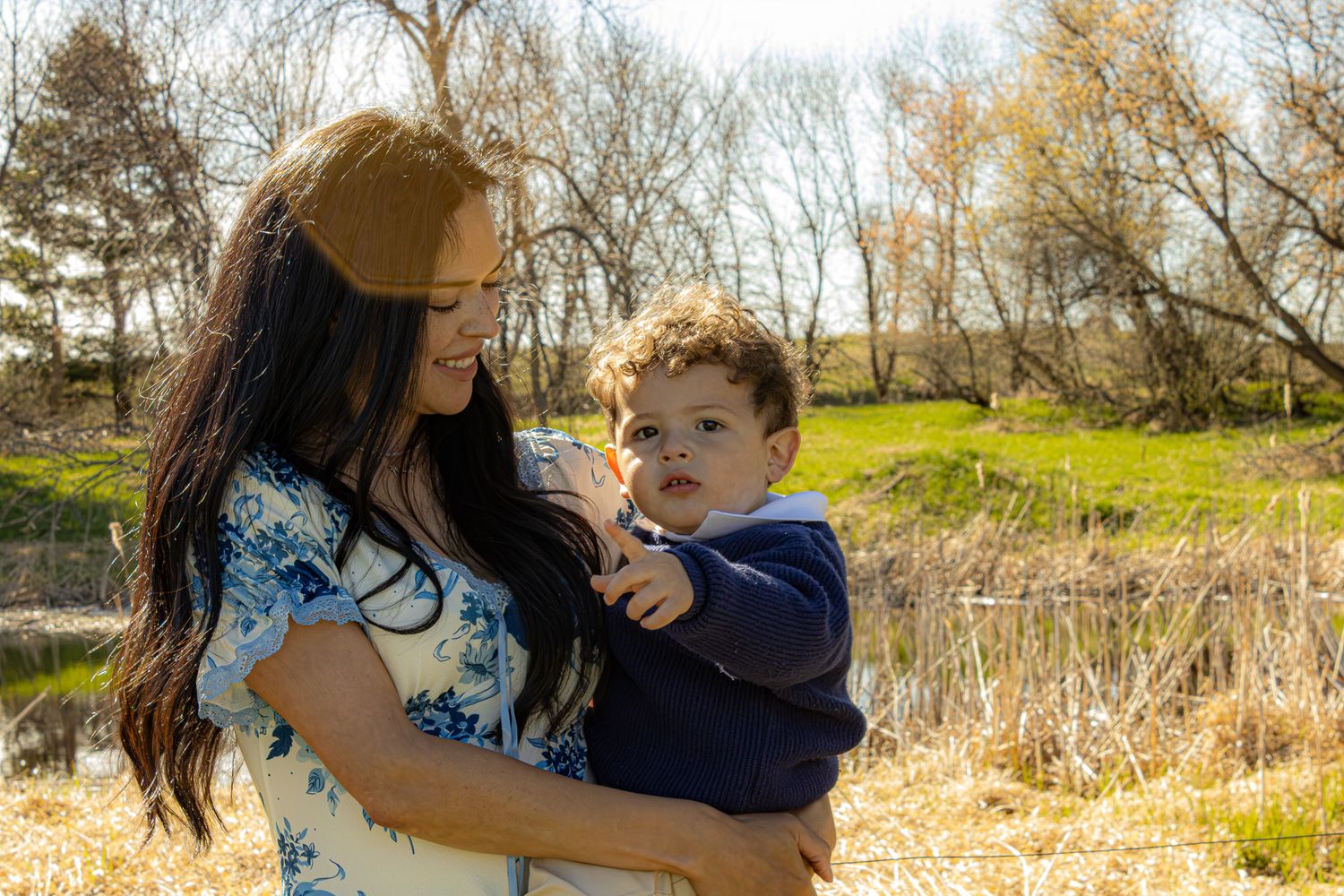 A mother holds her toddler son outdoors near a pond with bare trees and green grass in the background.