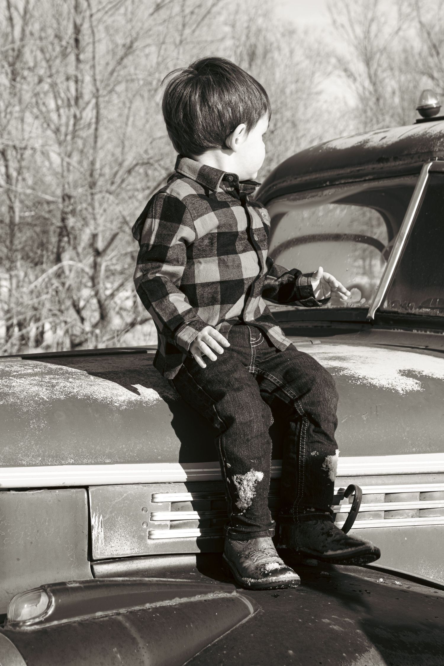 Young child in plaid shirt and jeans stands on hood of vintage car in black and white photograph.