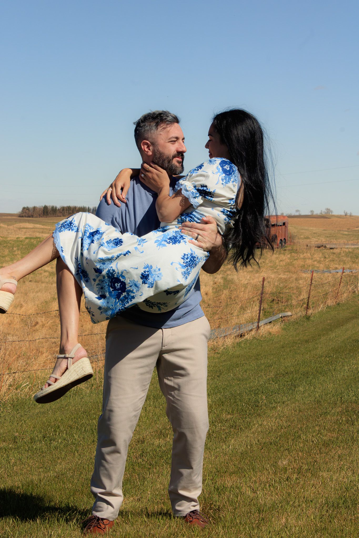 A man carries a woman in a floral dress outdoors in a sunny field, both smiling at each other romantically.