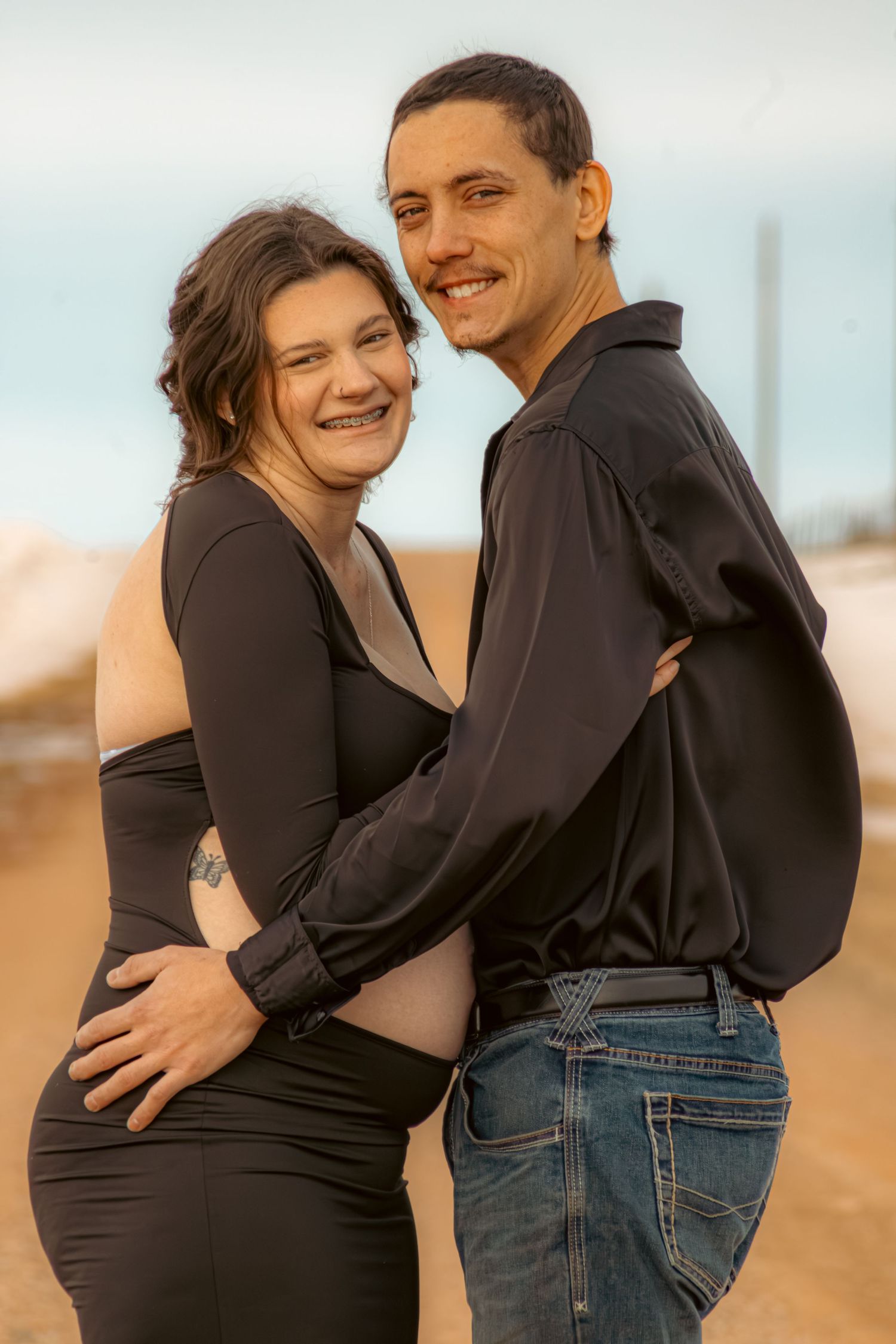 A couple shares an intimate embrace on a beach during a maternity photo shoot, wearing black and denim.