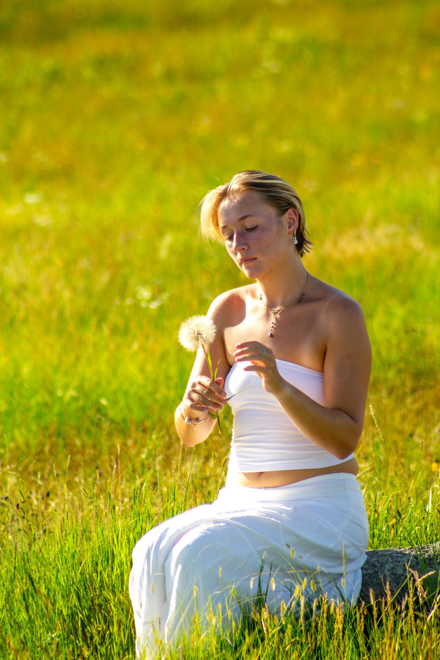 Young woman in white outfit sitting in green field holding dandelion flowers on sunny day.