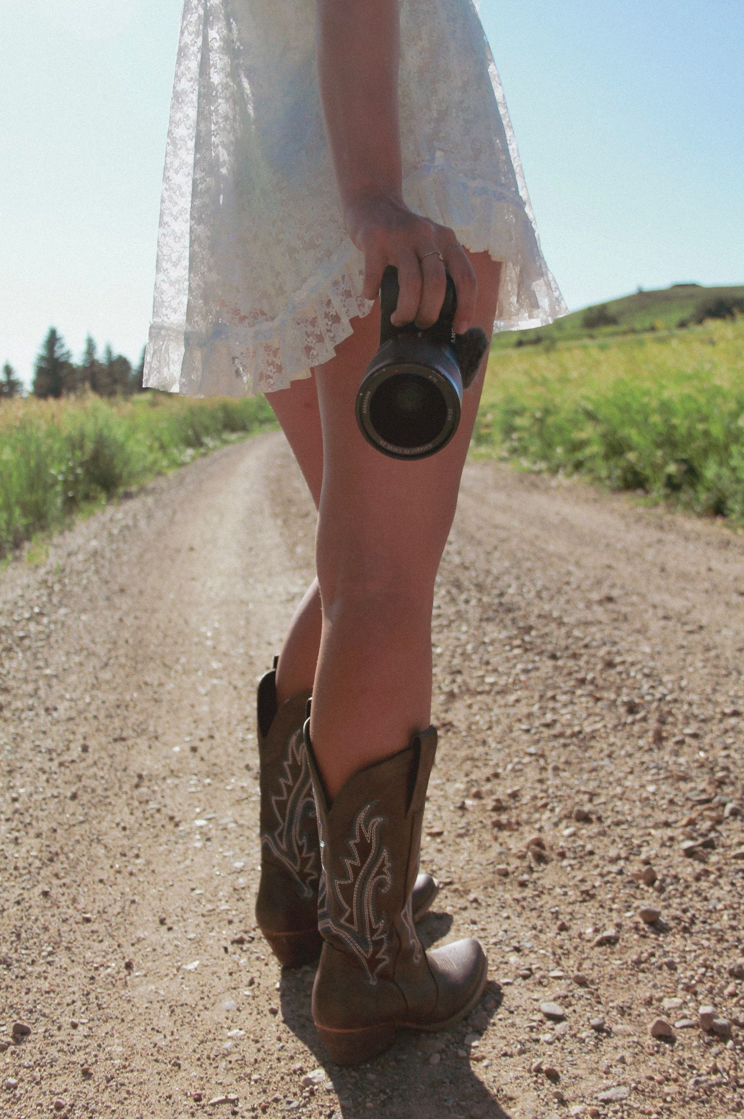 A pair of cowboy boots and white lace dress photographed on a dirt road with mountains in the distance.
