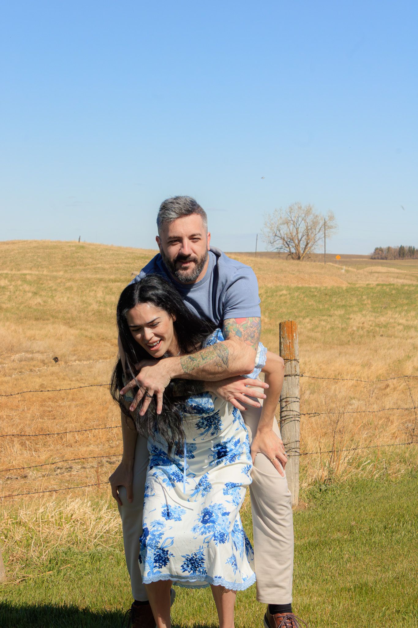 A couple embraces and smiles near a rustic fence in a sunny rural field, the man hugging the woman from behind.