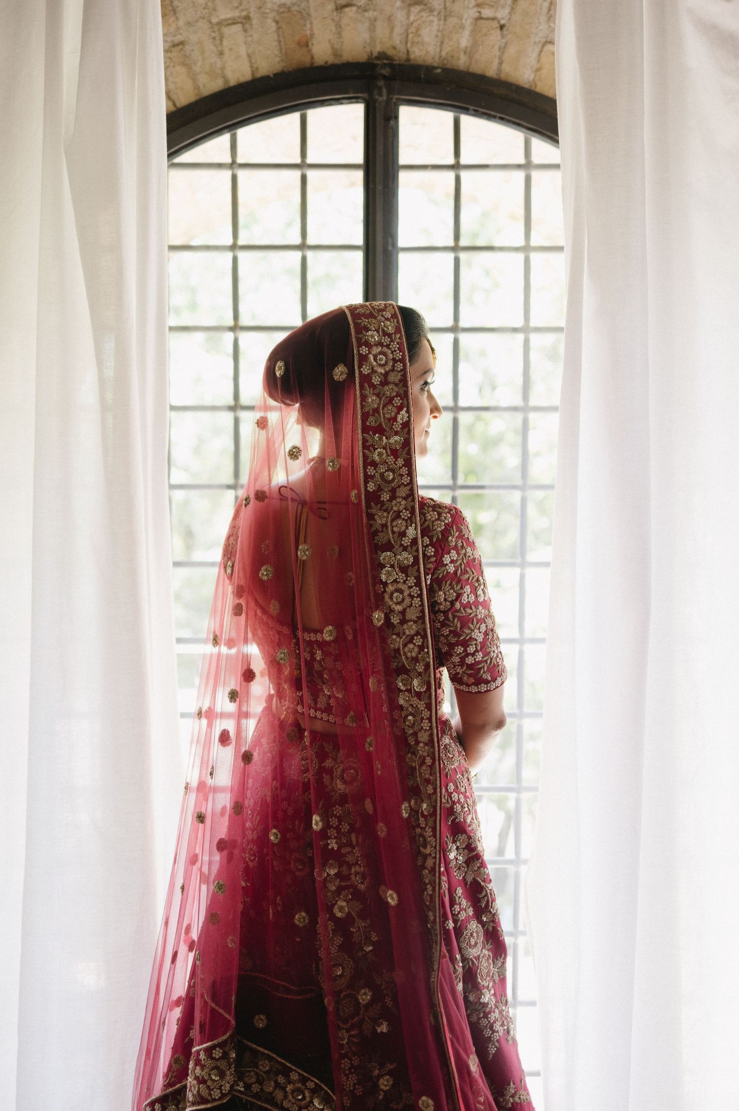 Traditional Indian bride in red and gold embroidered lehenga stands by arched window in elegant bridal pose.