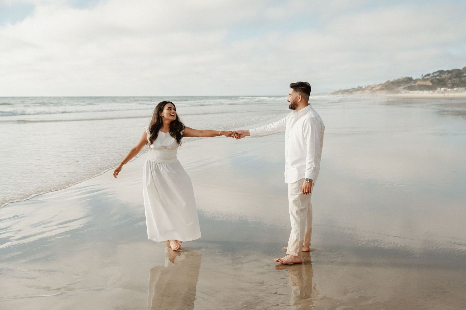 A couple in white outfits walks hand in hand along a scenic beach at sunset.