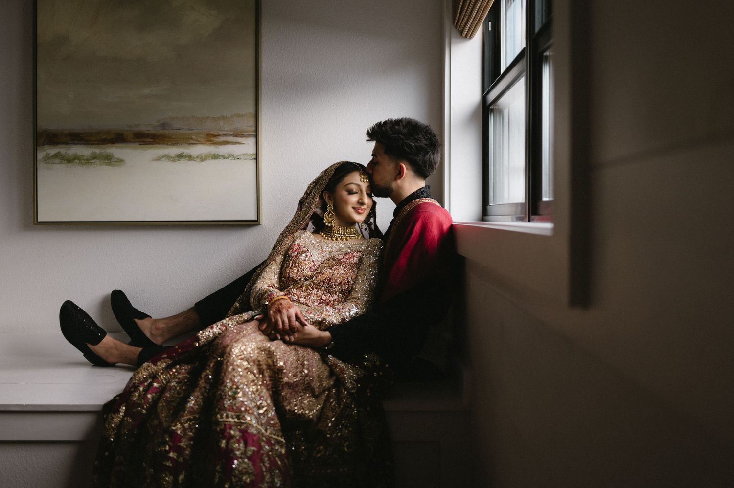 A couple in traditional Indian wedding attire sits together in an intimate indoor setting with soft natural light.