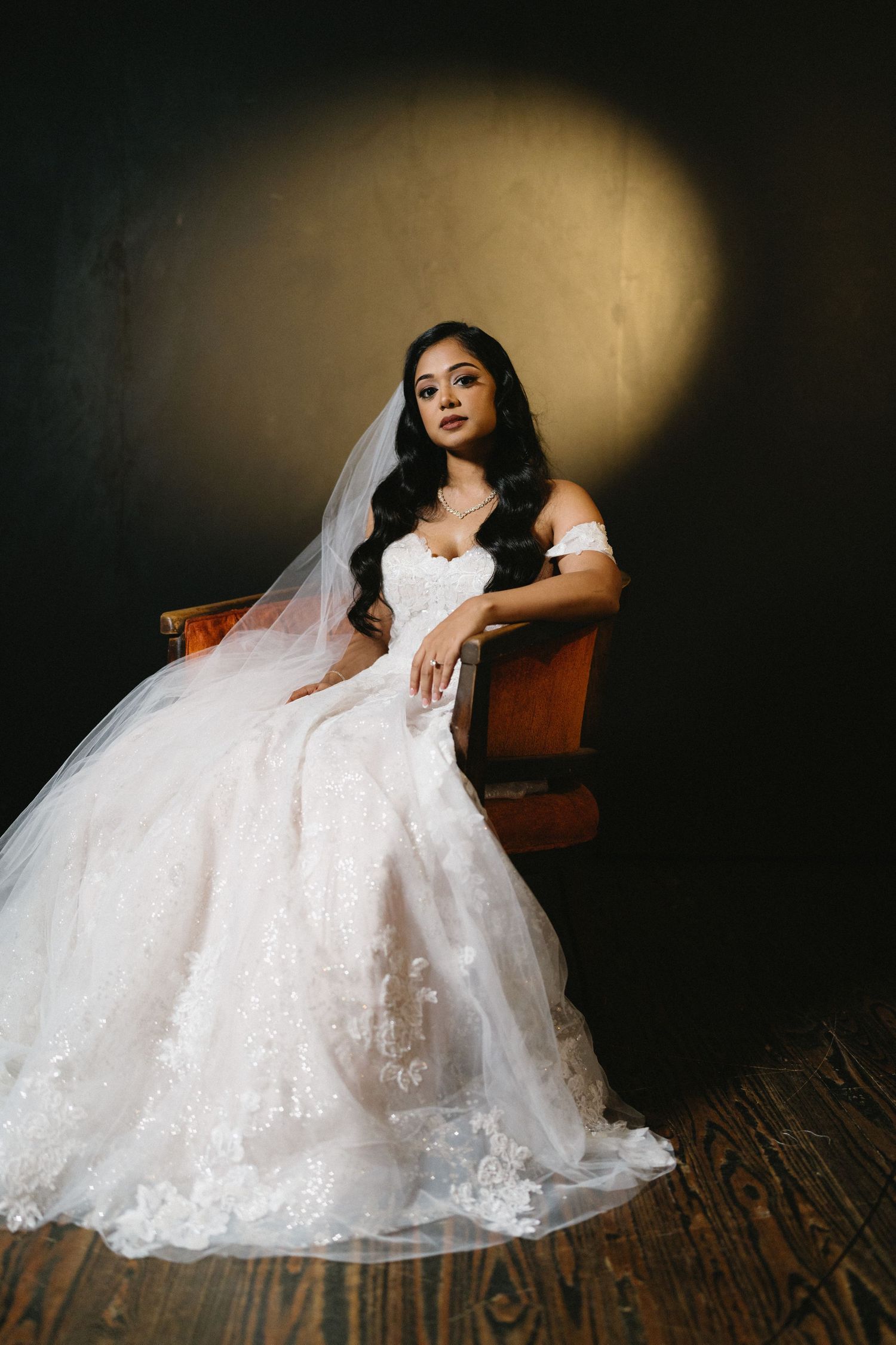 A bride in a white lace wedding gown poses elegantly on a wooden floor against a dark dramatic background.