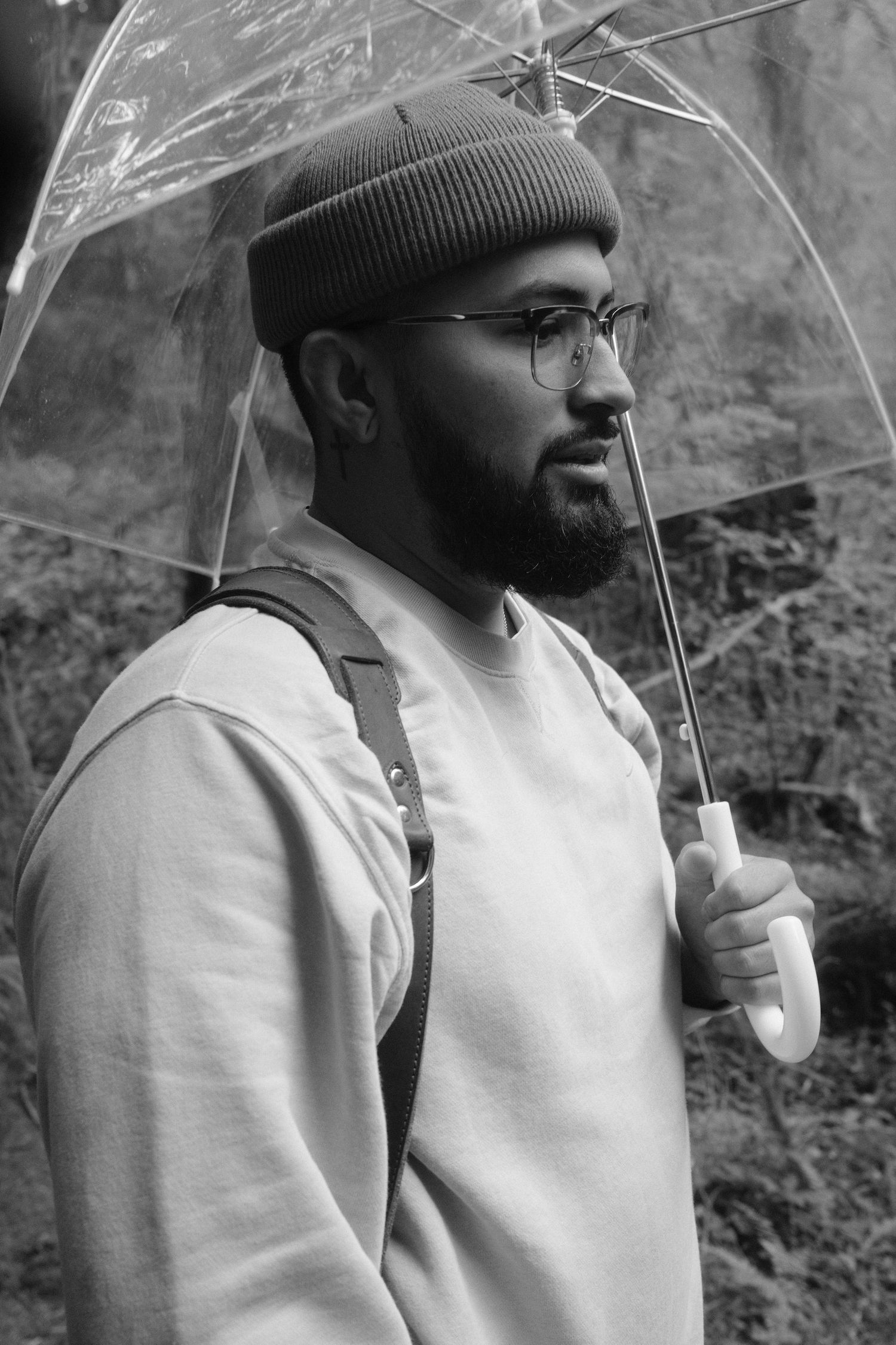 Black and white portrait of Jonny with glasses and a beanie holding a clear umbrella.