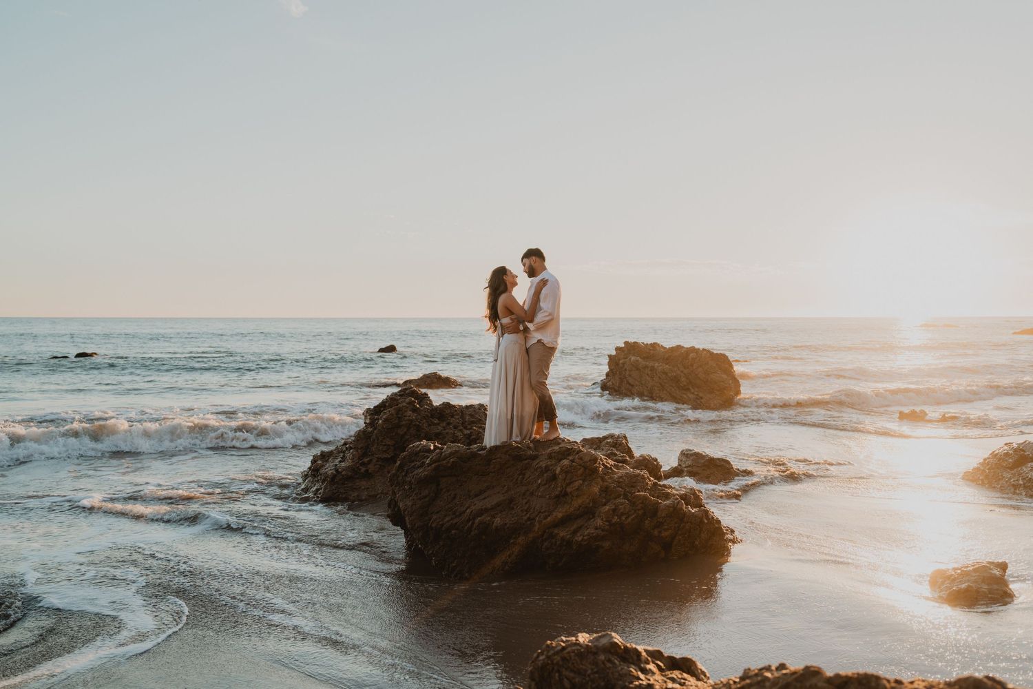 A couple stands on coastal rocks during golden hour as ocean waves crash against the shore at sunset.