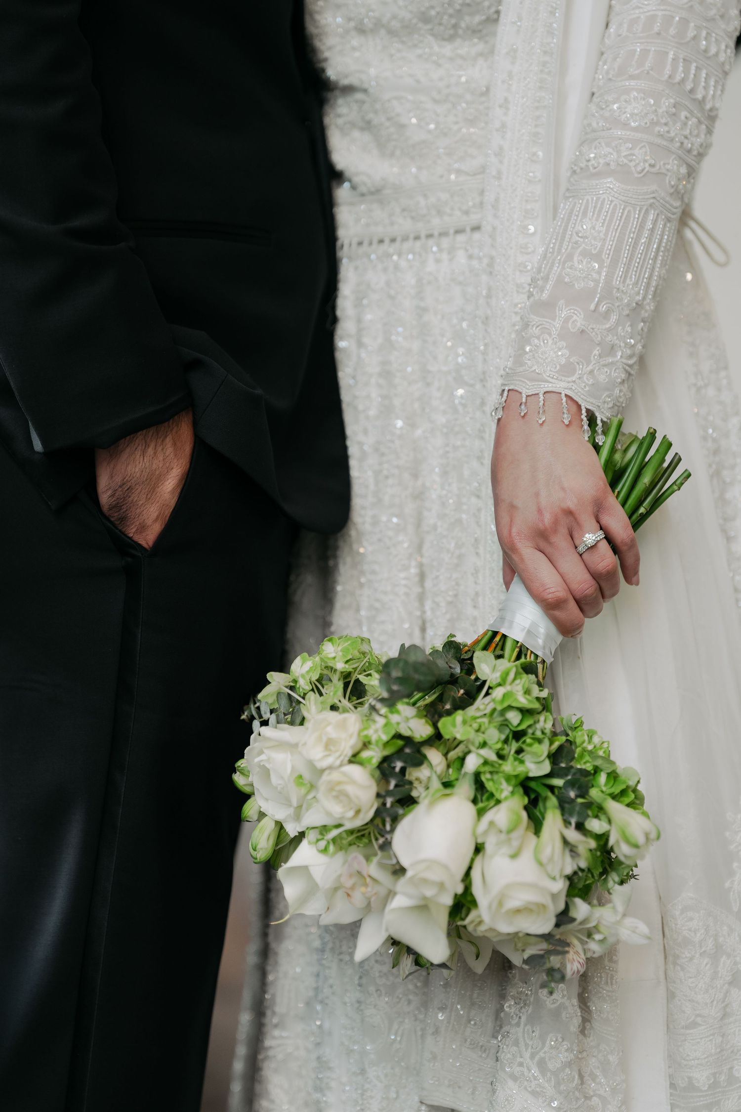 Close-up of couple in wedding attire holding white and green floral bouquet against lace wedding dress.
