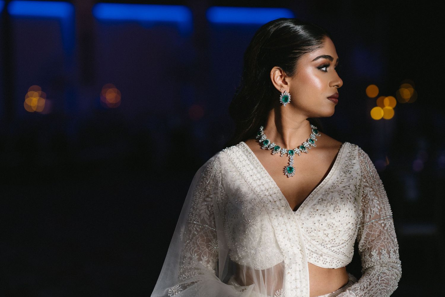 A person wearing an elegant white saree with intricate embroidery poses against a dark background with blue lighting.