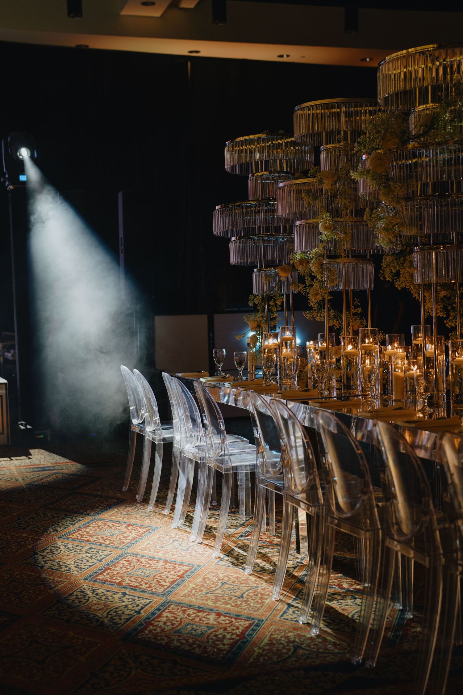Dramatic spotlight illuminates clear acrylic chairs and elegant table settings on a patterned rug in a dark event space.