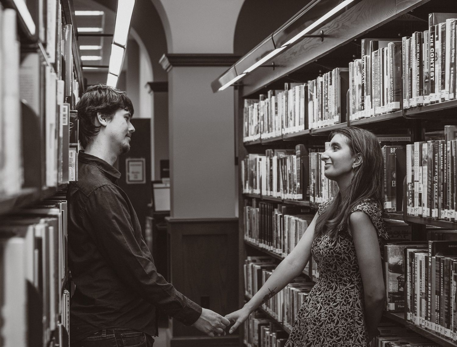 a black and white portrait of a couple holding hands and looking at each other in a library