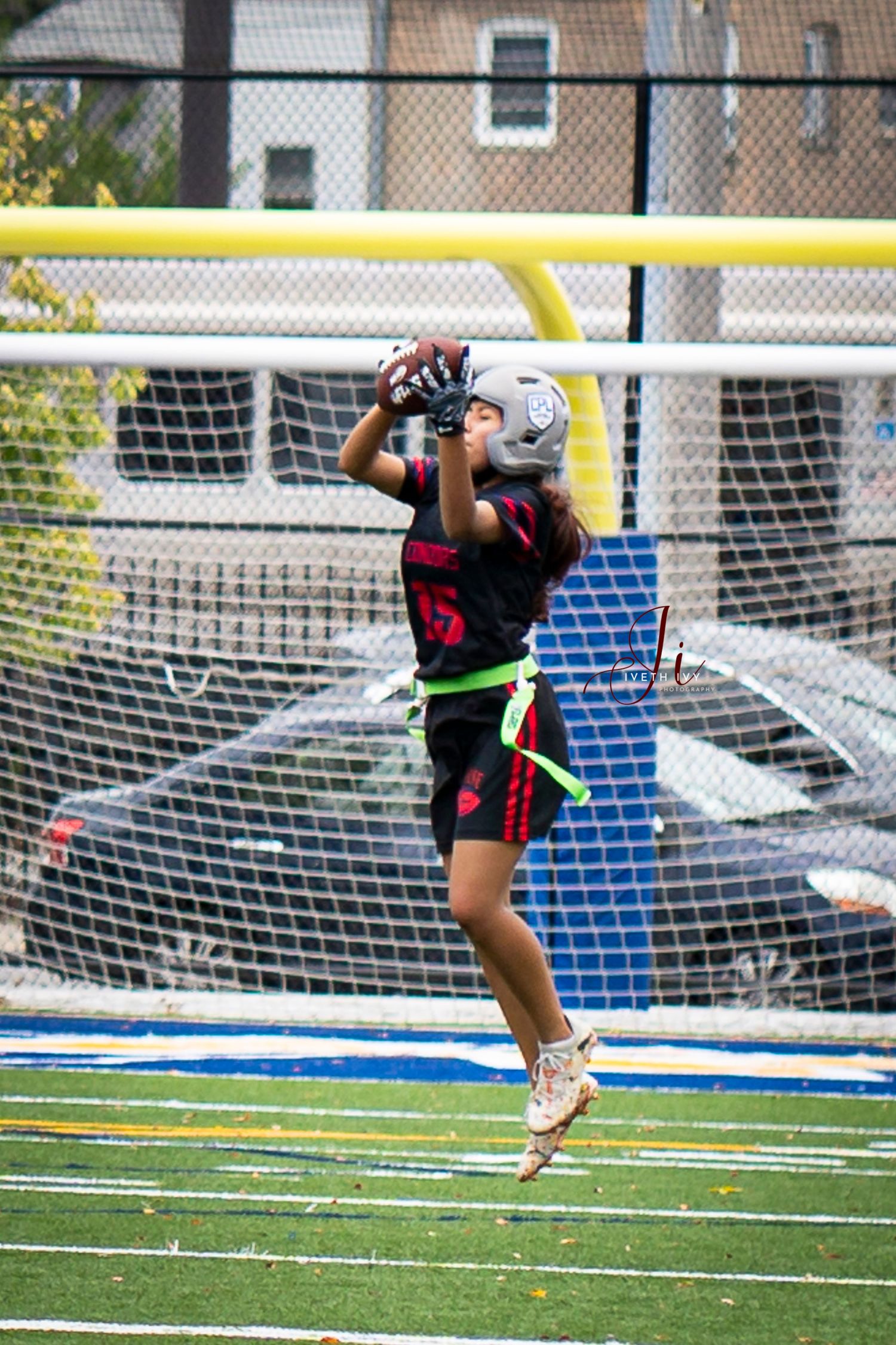 An athlete makes a dramatic diving save attempt during a soccer match on an outdoor turf field with a goal net visible.
