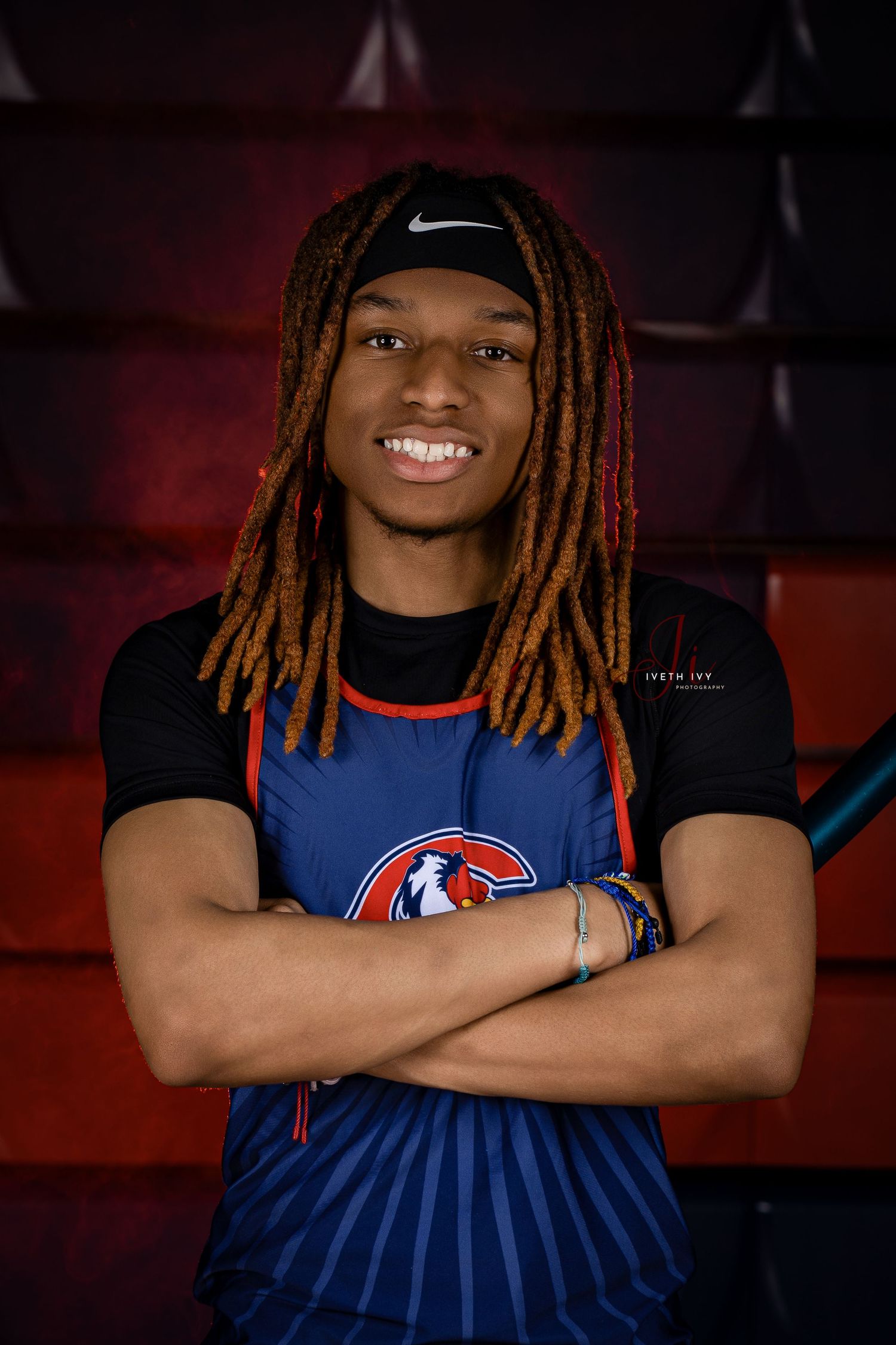 Athlete in blue uniform and black Nike cap with long dreadlocks smiles with arms crossed against red background.