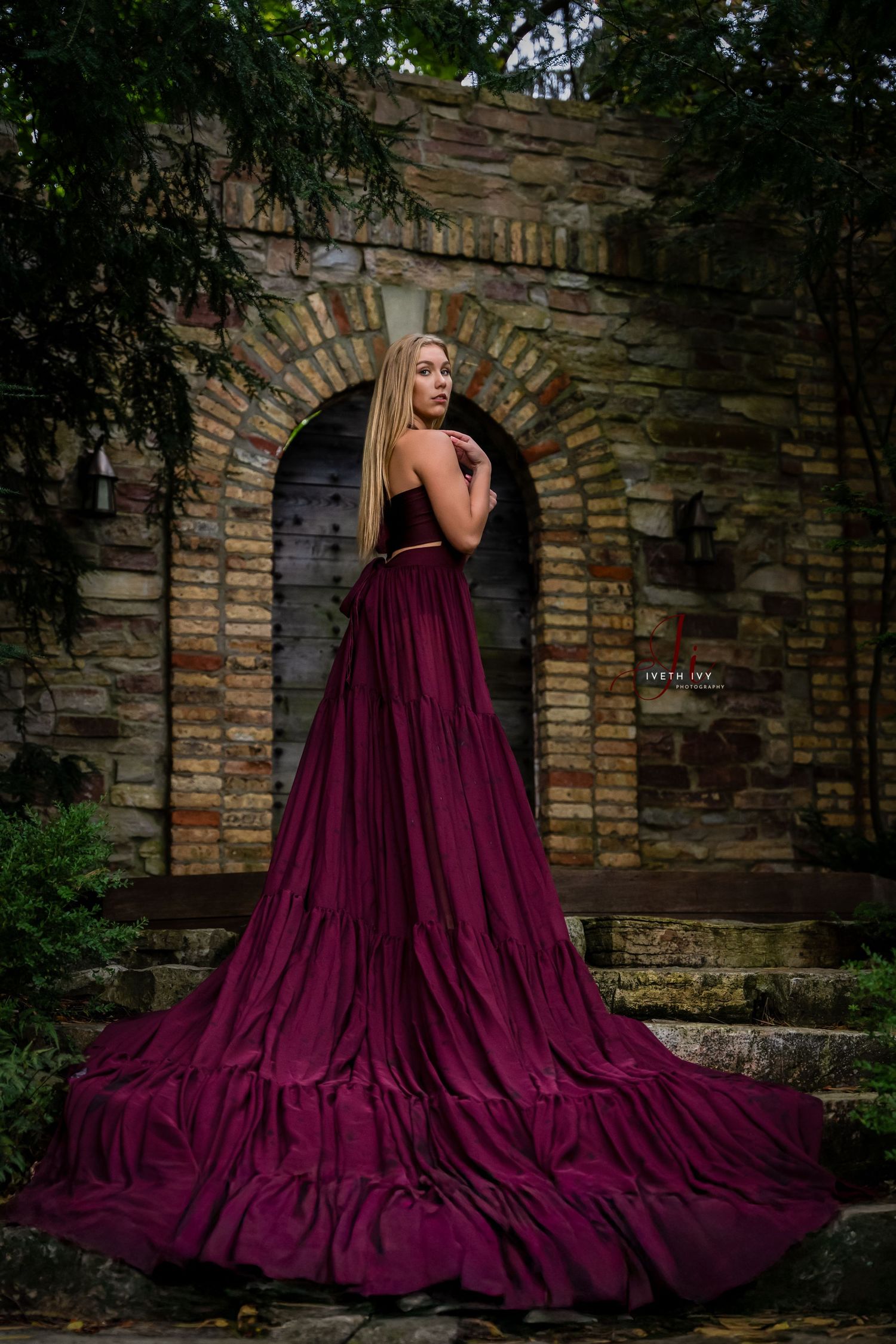 A dramatic burgundy ball gown with flowing train against a stone archway in a romantic garden setting.