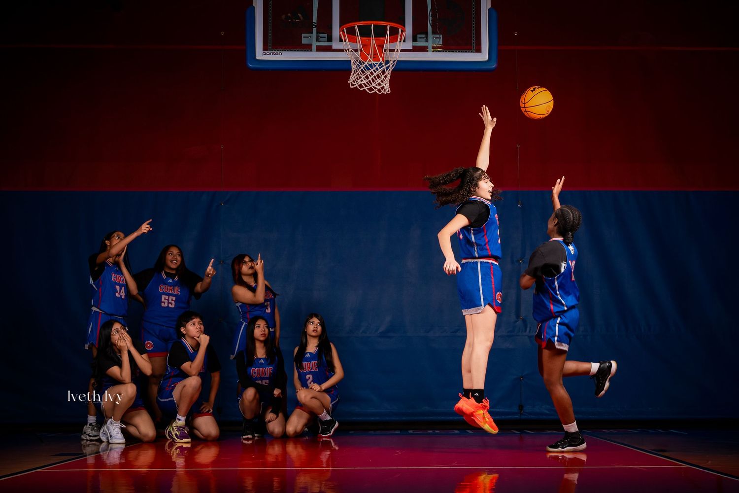 Basketball players in blue uniforms leap for the ball during an indoor game with dramatic lighting.