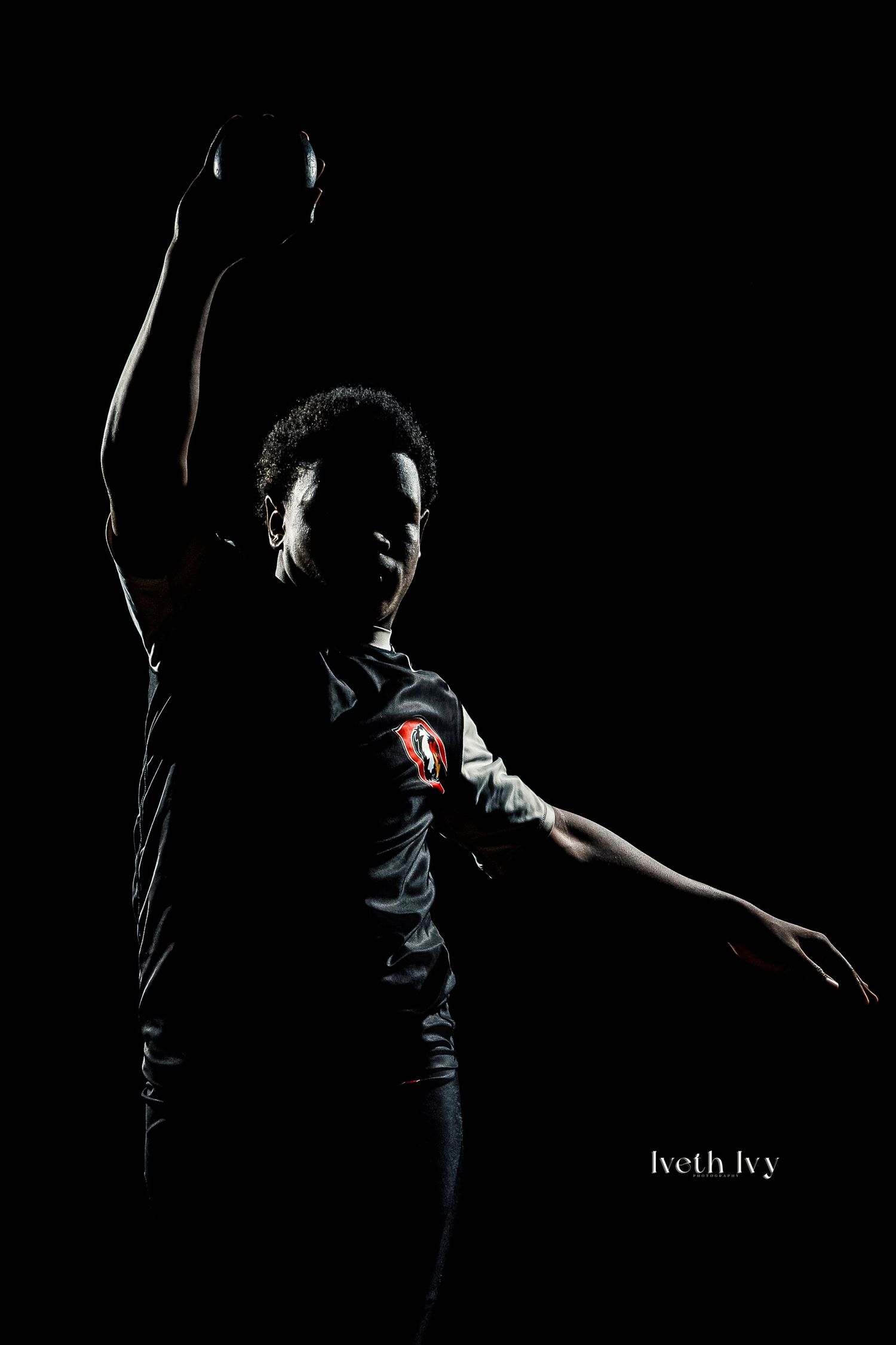 Silhouetted athlete holding a shot put ball overhead in dramatic low-key studio lighting, wearing a dark sports jersey.
