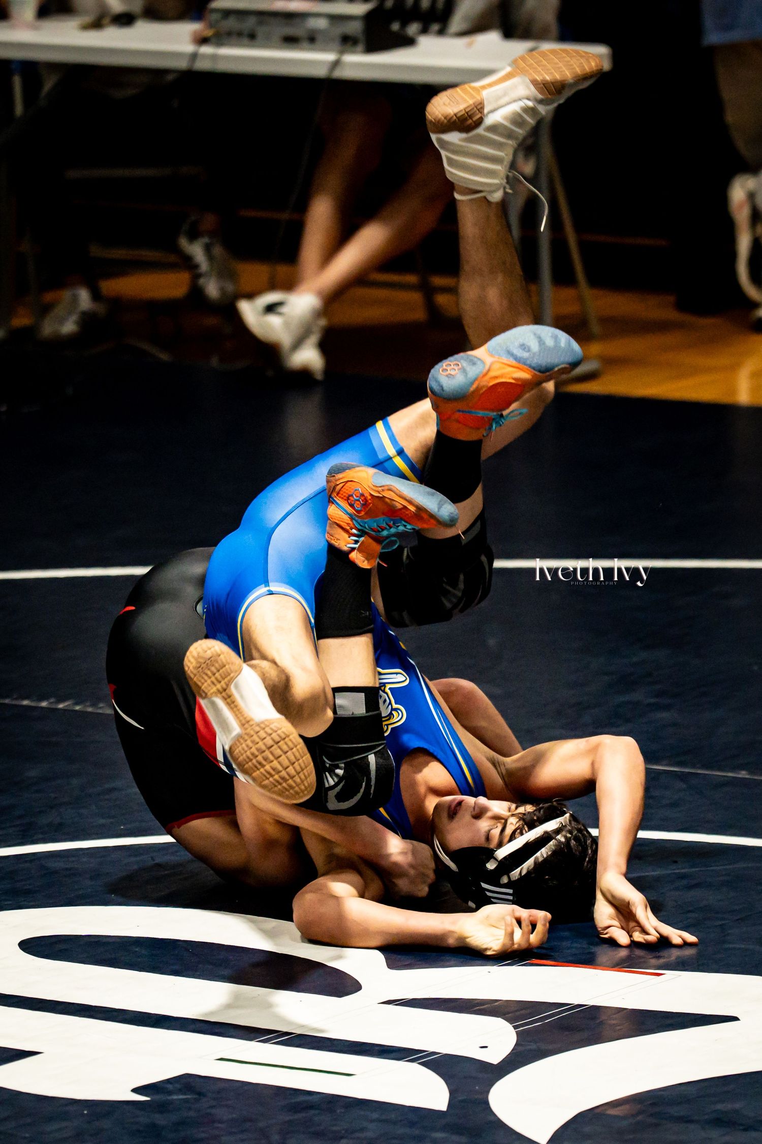 Two wrestlers compete on a blue mat during an intense wrestling match with one athlete attempting a pin maneuver.