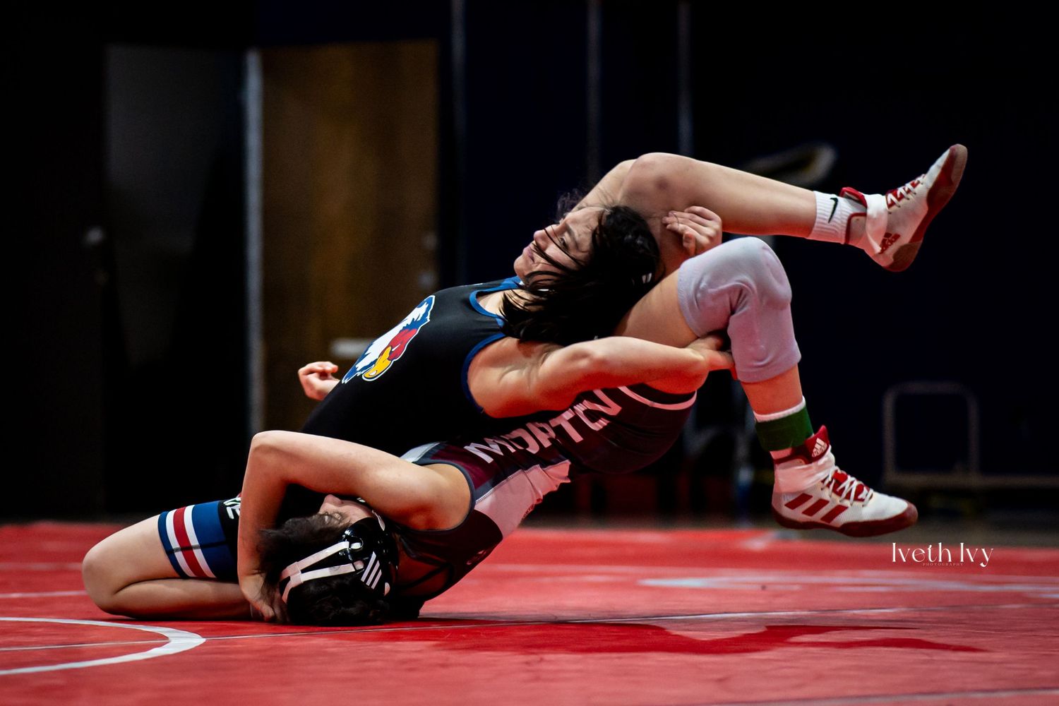 Wrestlers compete in a dynamic match on a red wrestling mat with spectators watching in the background.