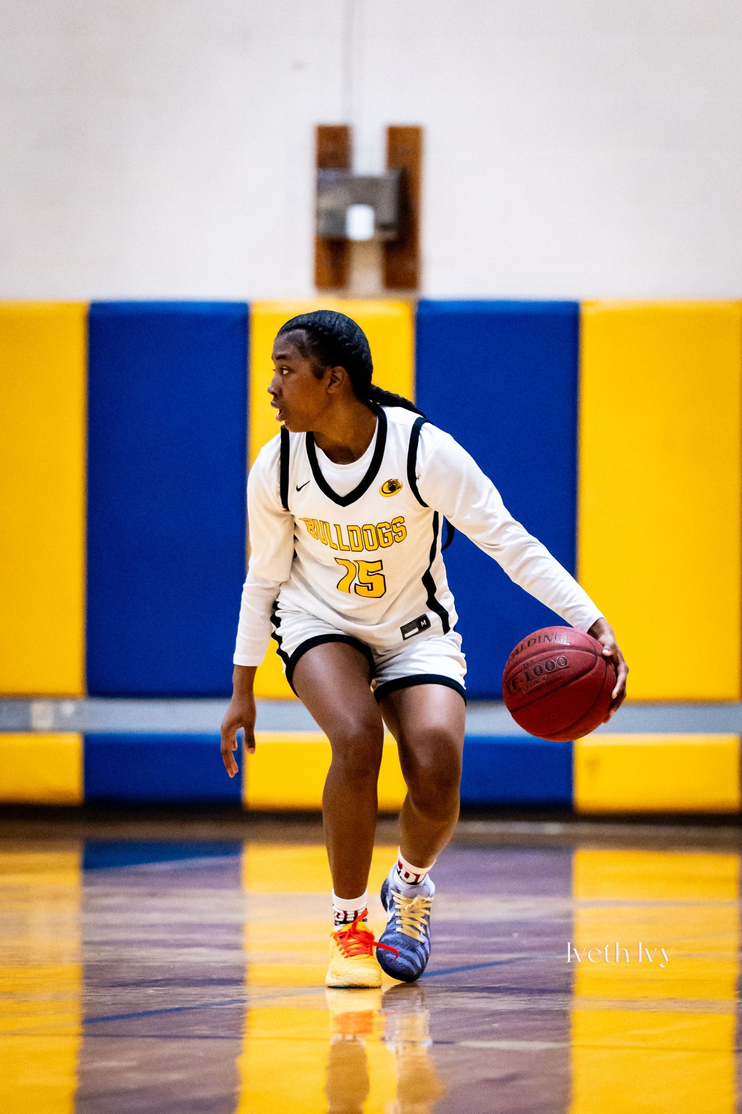 Female basketball player in Bulldogs #75 jersey dribbling on indoor court with blue and yellow walls.