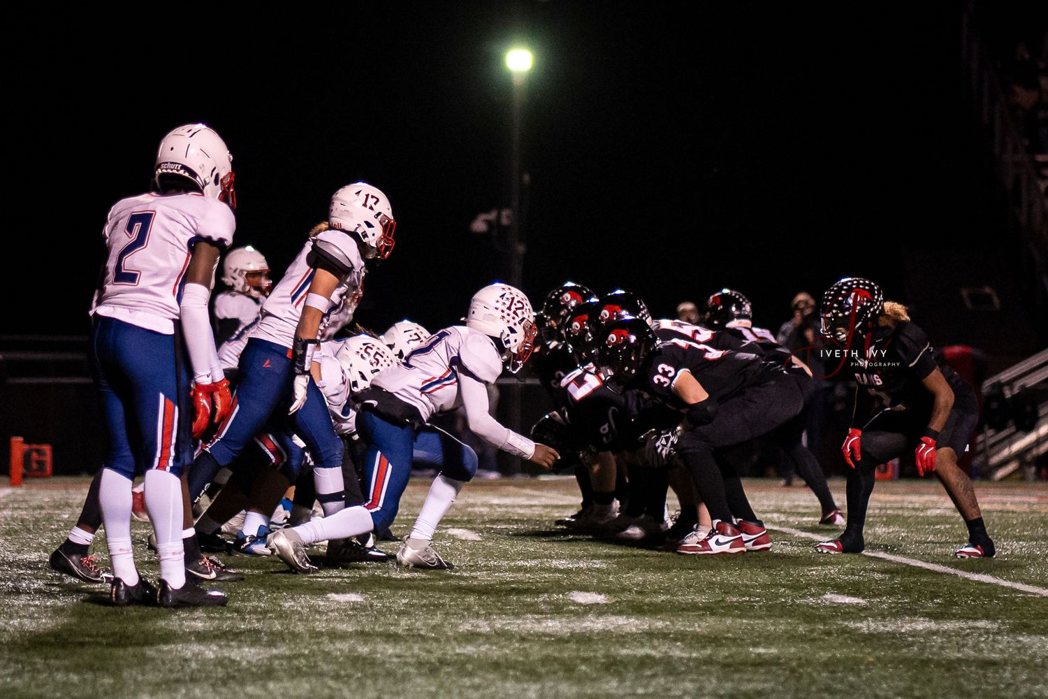 High school football players line up at the line of scrimmage under stadium lights during a nighttime game.
