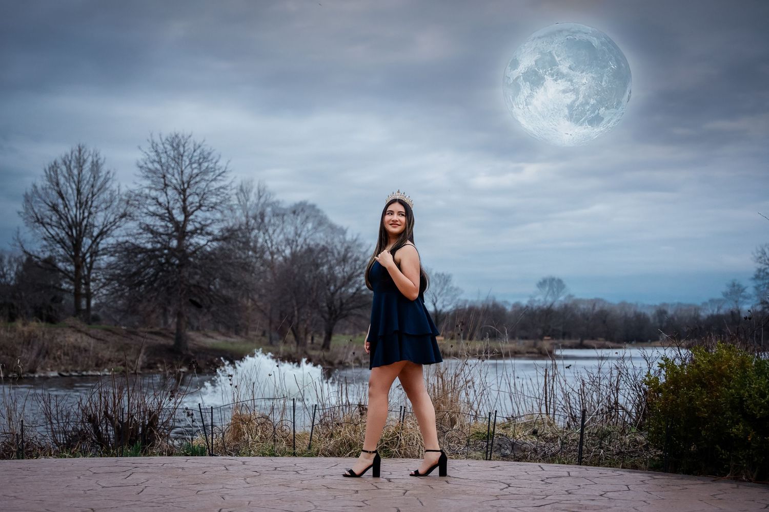 A figure in a short black dress poses by a lake under a dramatic full moon in a moody evening landscape.