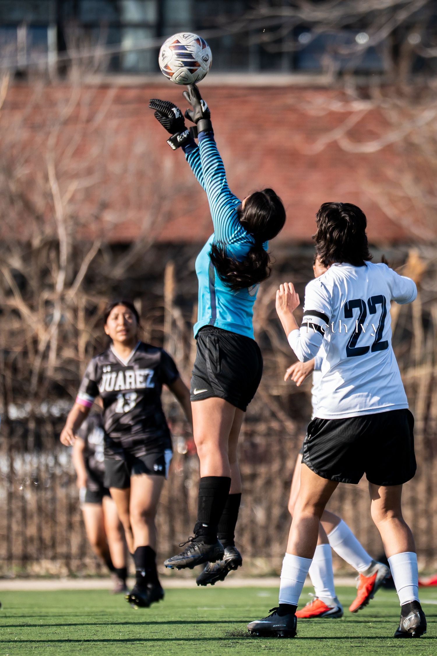Female soccer goalkeeper in blue jumps to block ball during competitive match with player number 22 nearby.