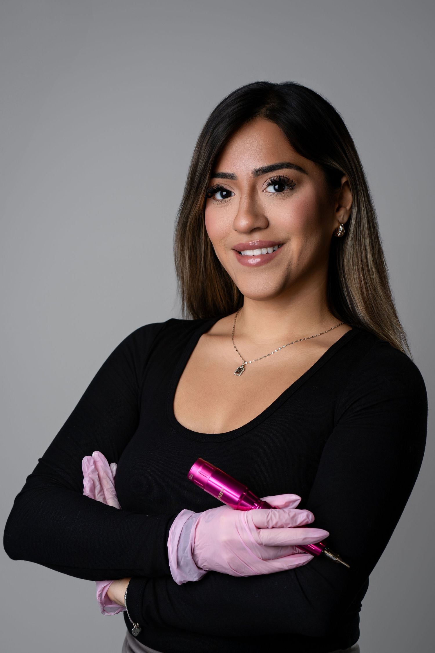 A smiling woman in a black top wearing pink gloves holds a pink tattoo machine with arms crossed against a gray background.