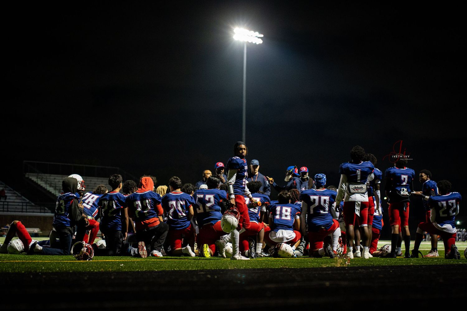 Football team practices under bright stadium lights at night on a grass field wearing red and blue uniforms.