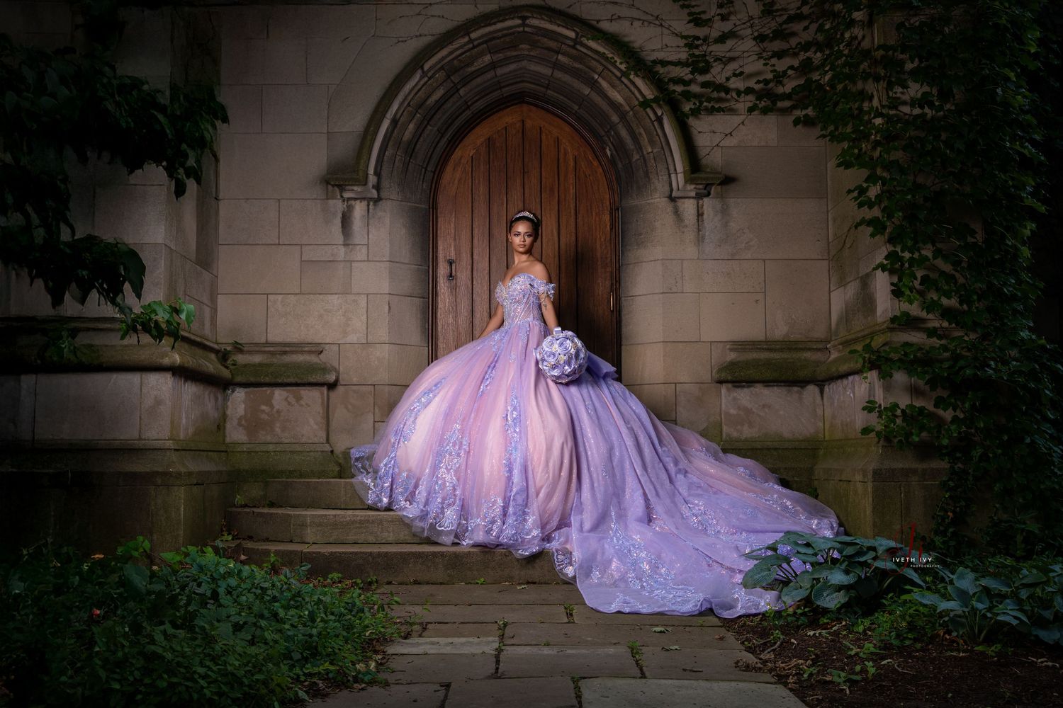 Purple ball gown with dramatic train flows across stone steps in romantic garden setting at dusk.