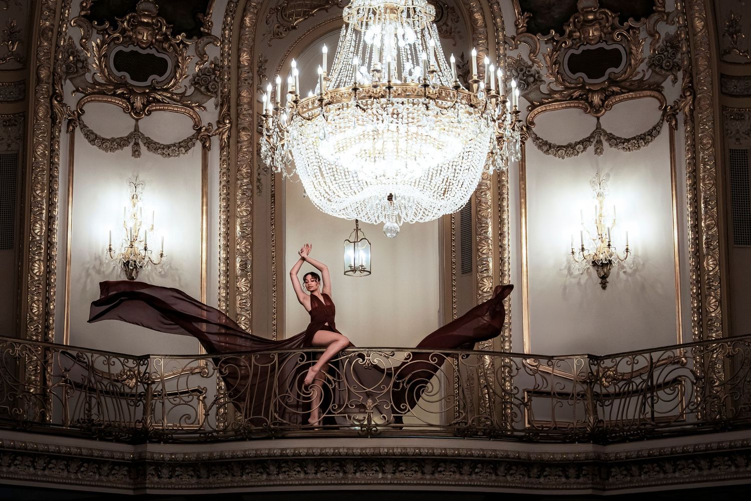 Elegant figure in red dress poses gracefully on ornate staircase beneath crystal chandelier in baroque palace interior.