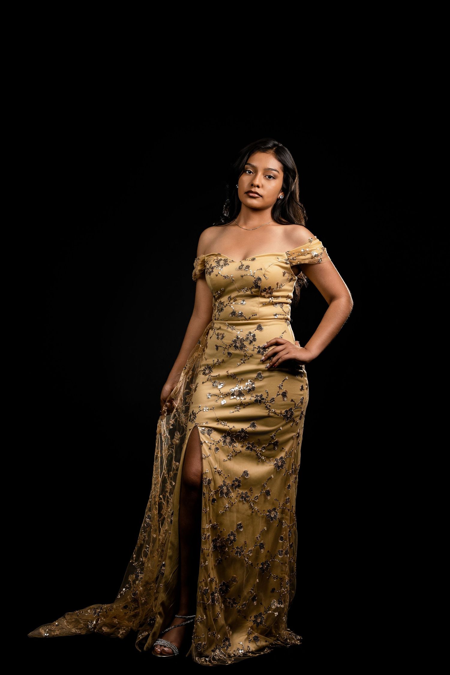 Woman in gold floral off-shoulder gown with slit poses elegantly against black background.