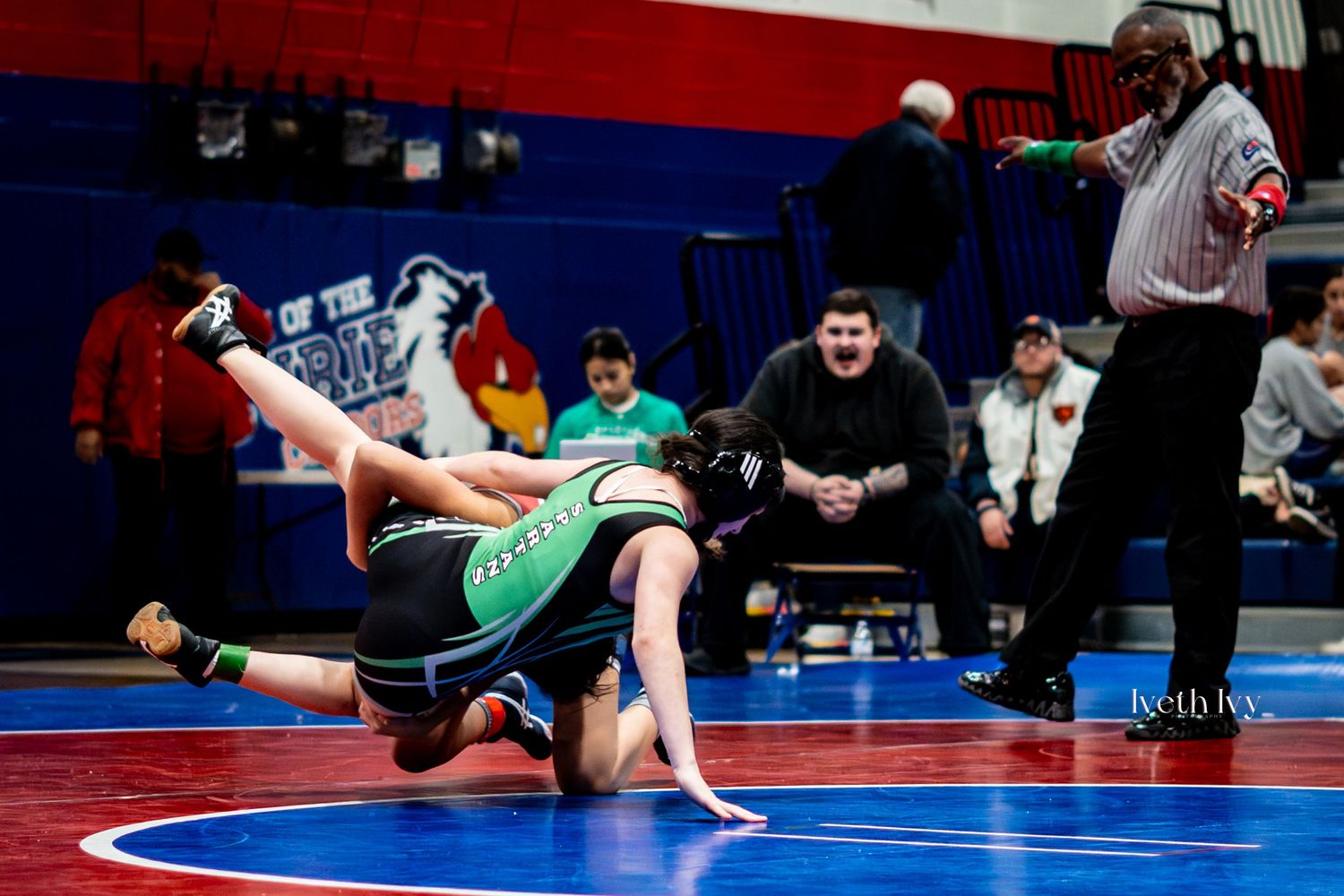 Athletes engage in a wrestling match on a blue and red competition mat under arena lighting.