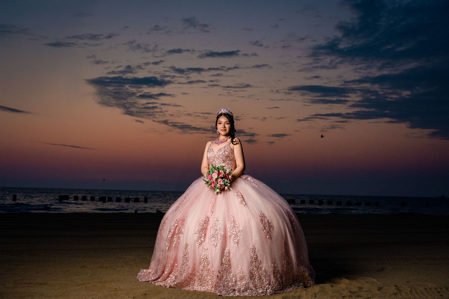 A pink quinceañera dress silhouetted against a dramatic sunset beach backdrop with clouds and ocean pier.