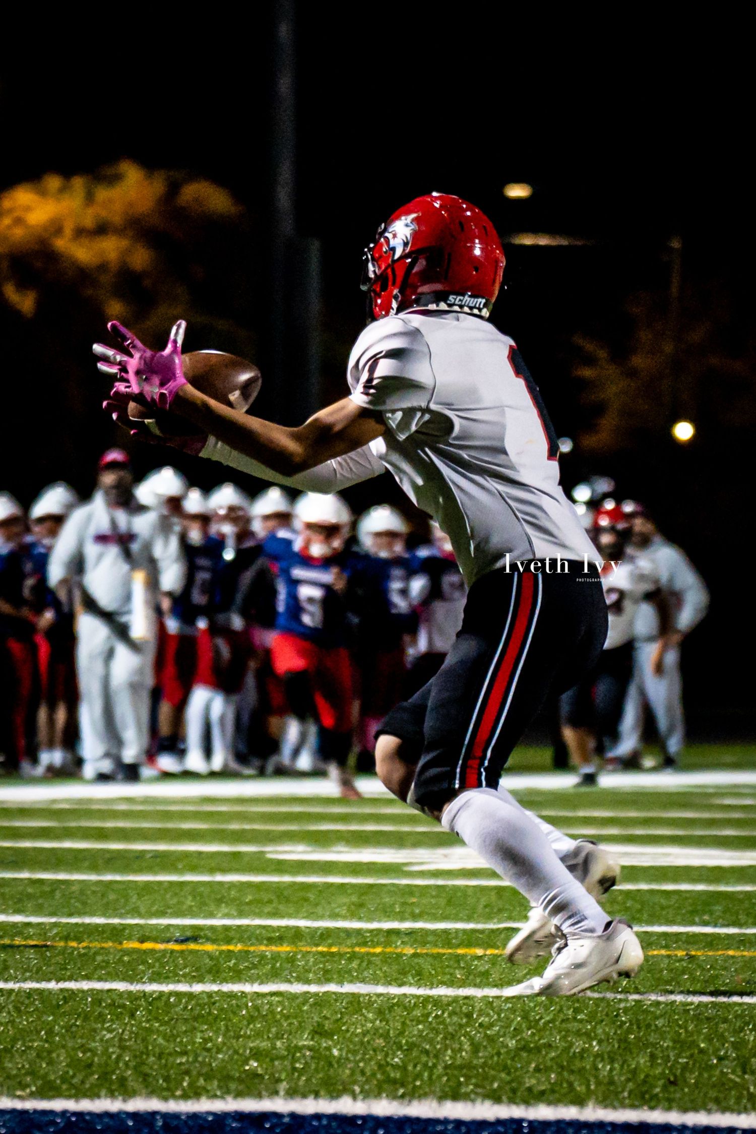 Football player in red helmet and white uniform reaches out to catch the ball during night game.