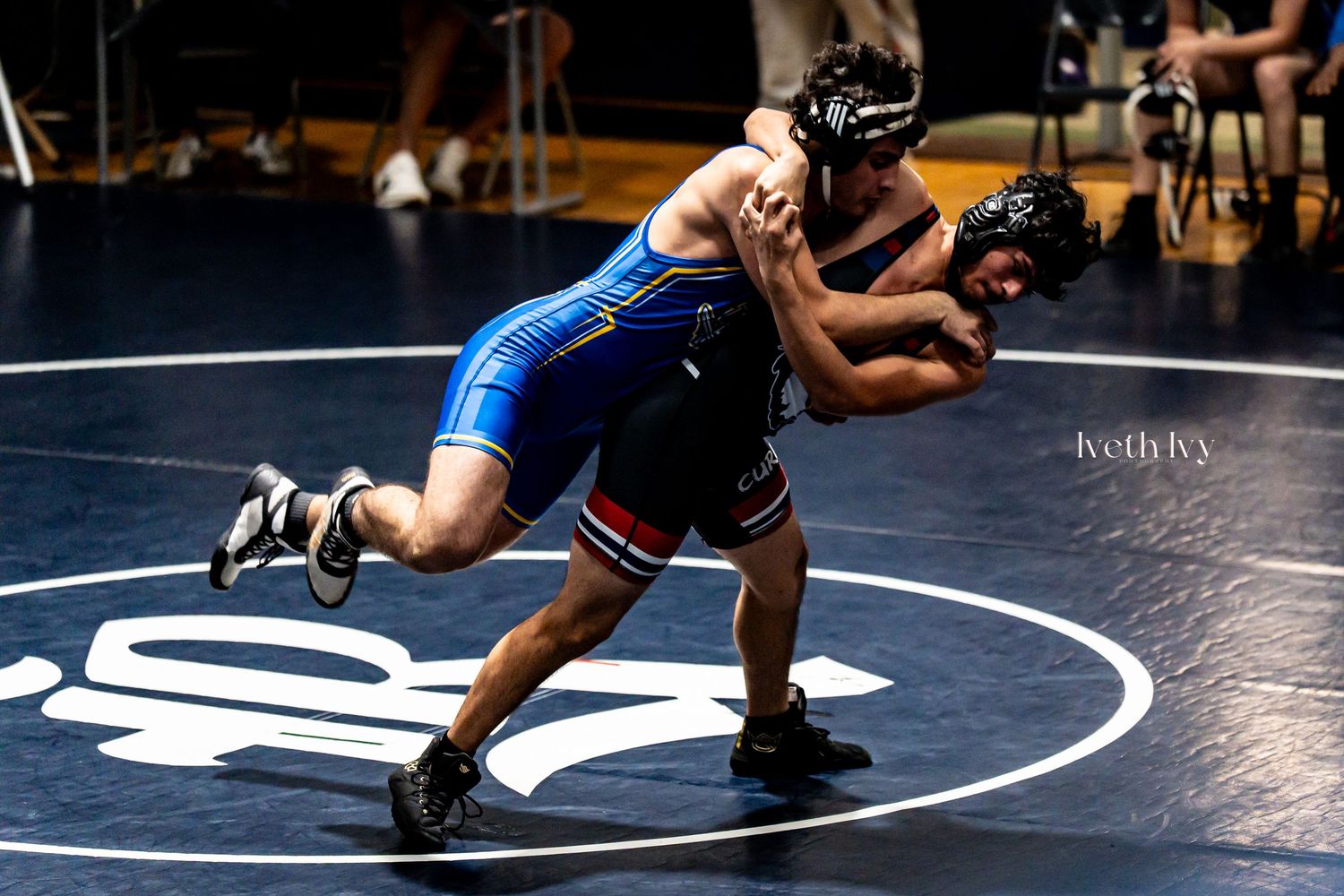 Two wrestlers compete in a high school wrestling match on a dark mat with dramatic lighting.