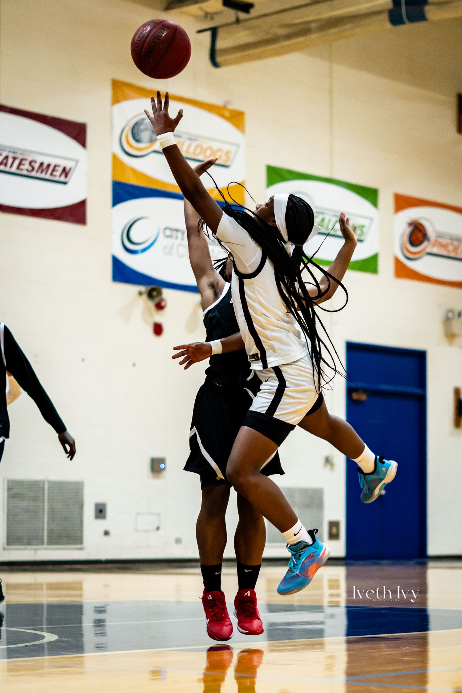 Two female basketball players jump for the ball during an indoor game, one reaching high to block or catch the shot.