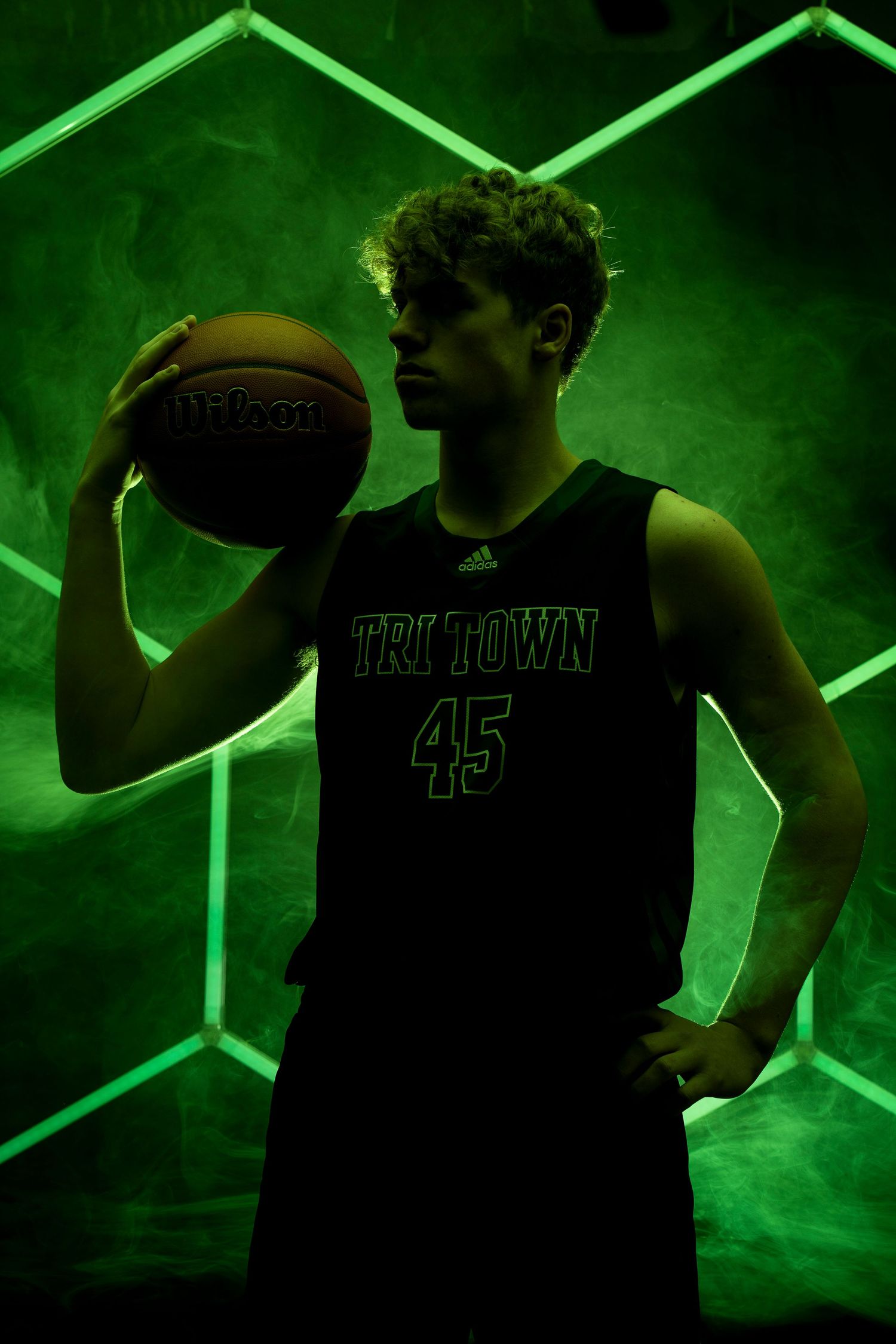 Basketball player in dark jersey poses with ball against green geometric background.