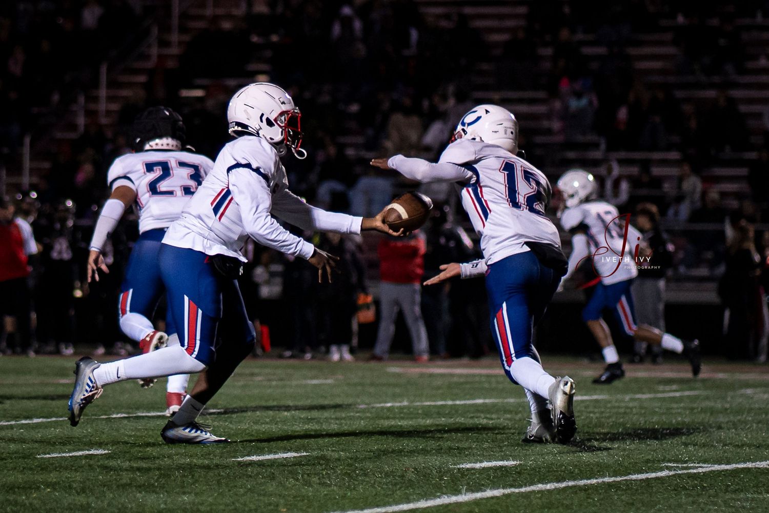 Football players in white uniforms with blue accents move across a dark field during a nighttime game.