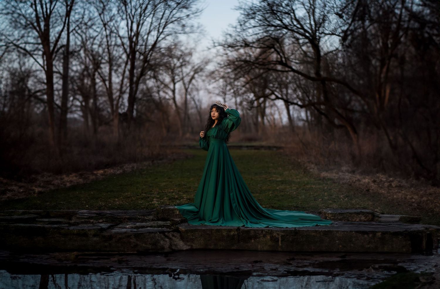 Ethereal emerald green gown photographed at dusk by still water with bare trees in background.