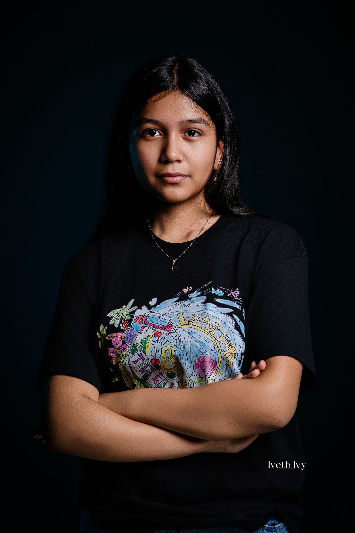 Young woman in graphic black tee poses confidently with arms crossed against dark teal studio background.