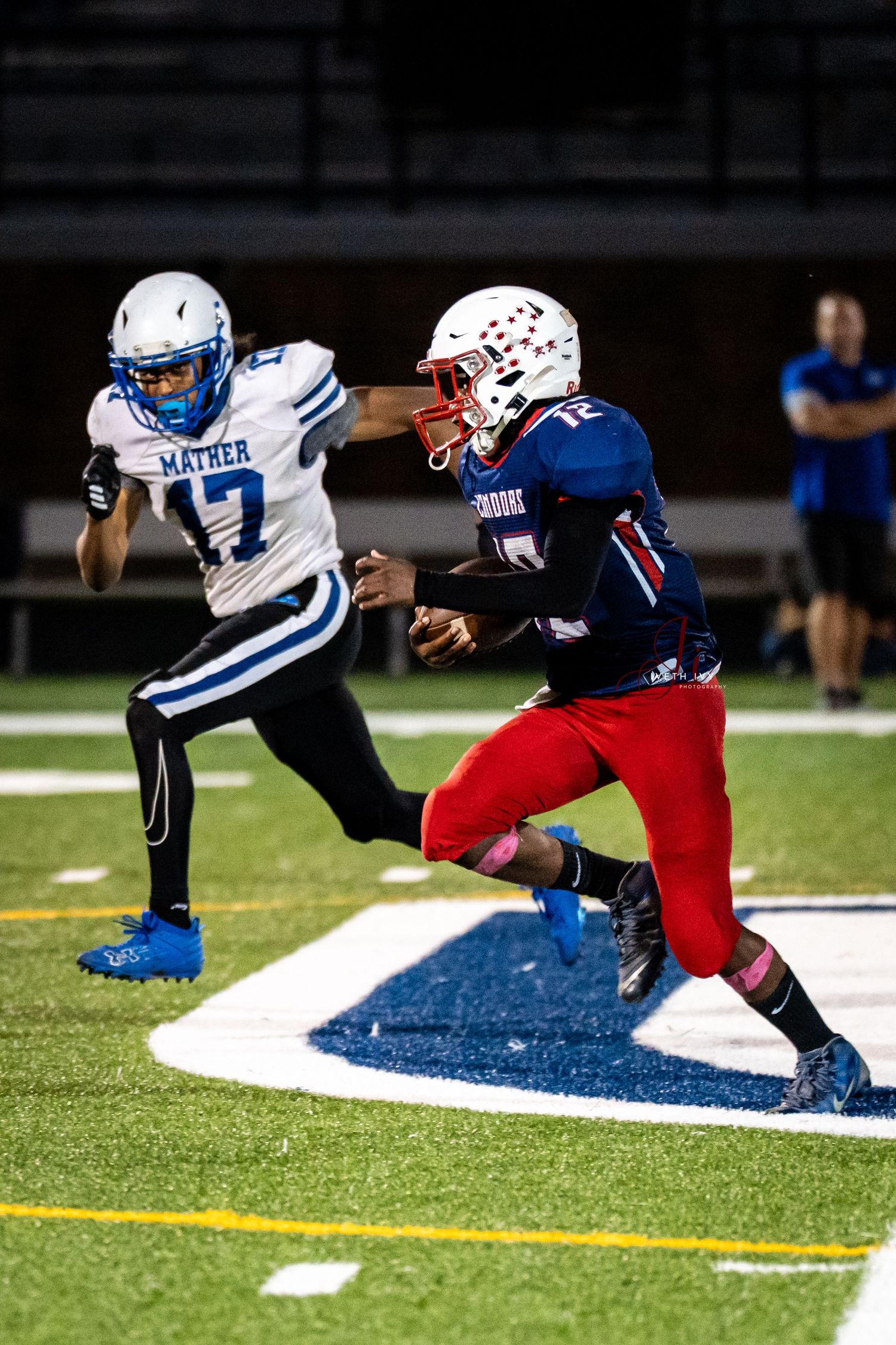 Football player in blue jersey diving to tackle opponent in red uniform during nighttime game on turf field.