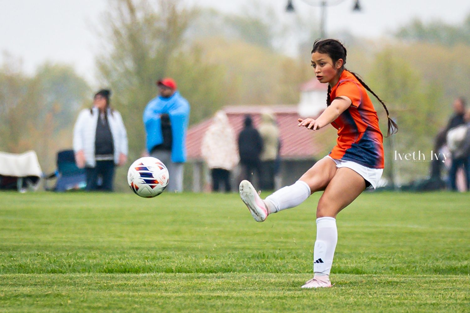 Young female soccer player in orange jersey kicking ball during outdoor match on green field.