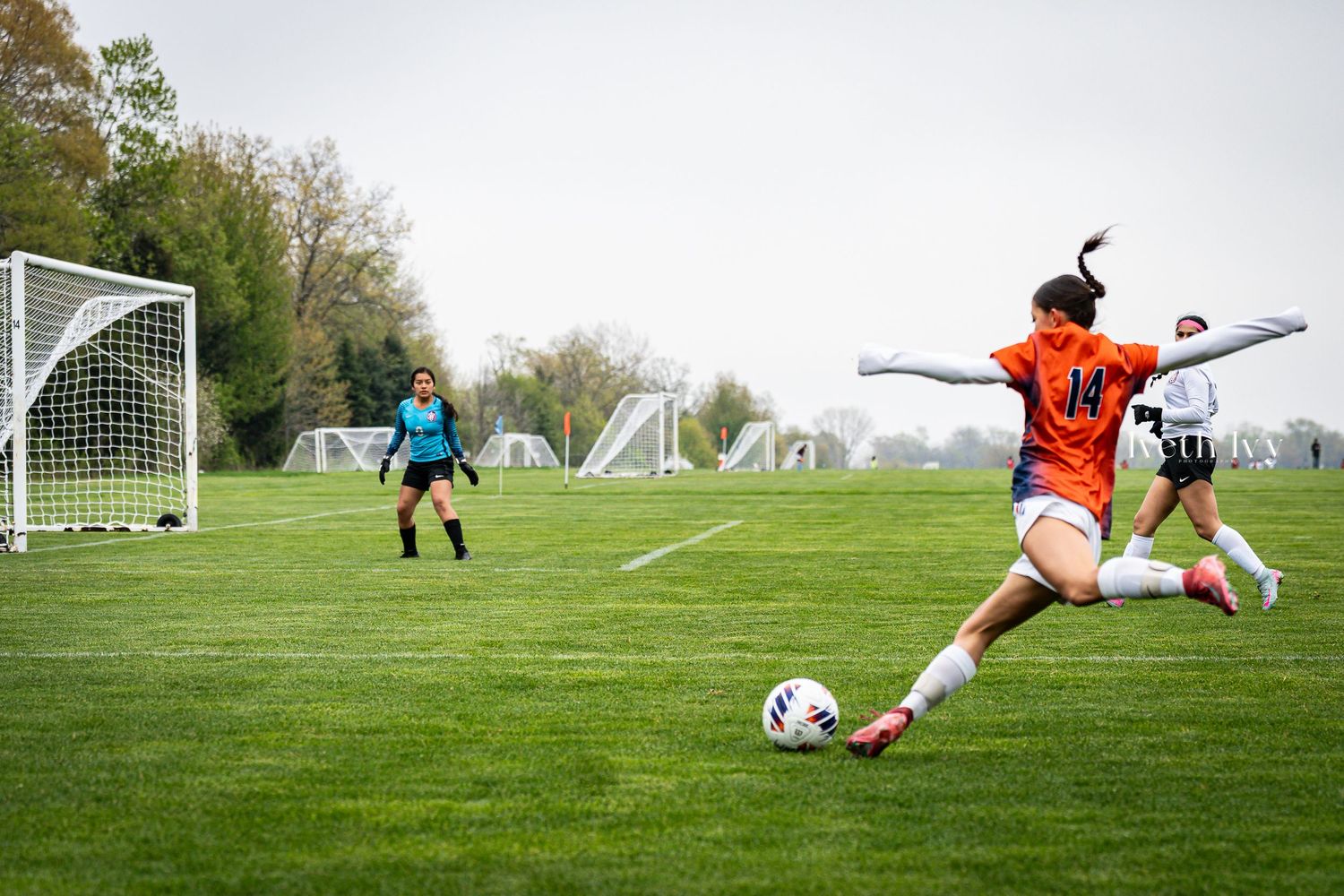 Female soccer player in orange jersey kicks ball toward goal during a competitive match on a green field.